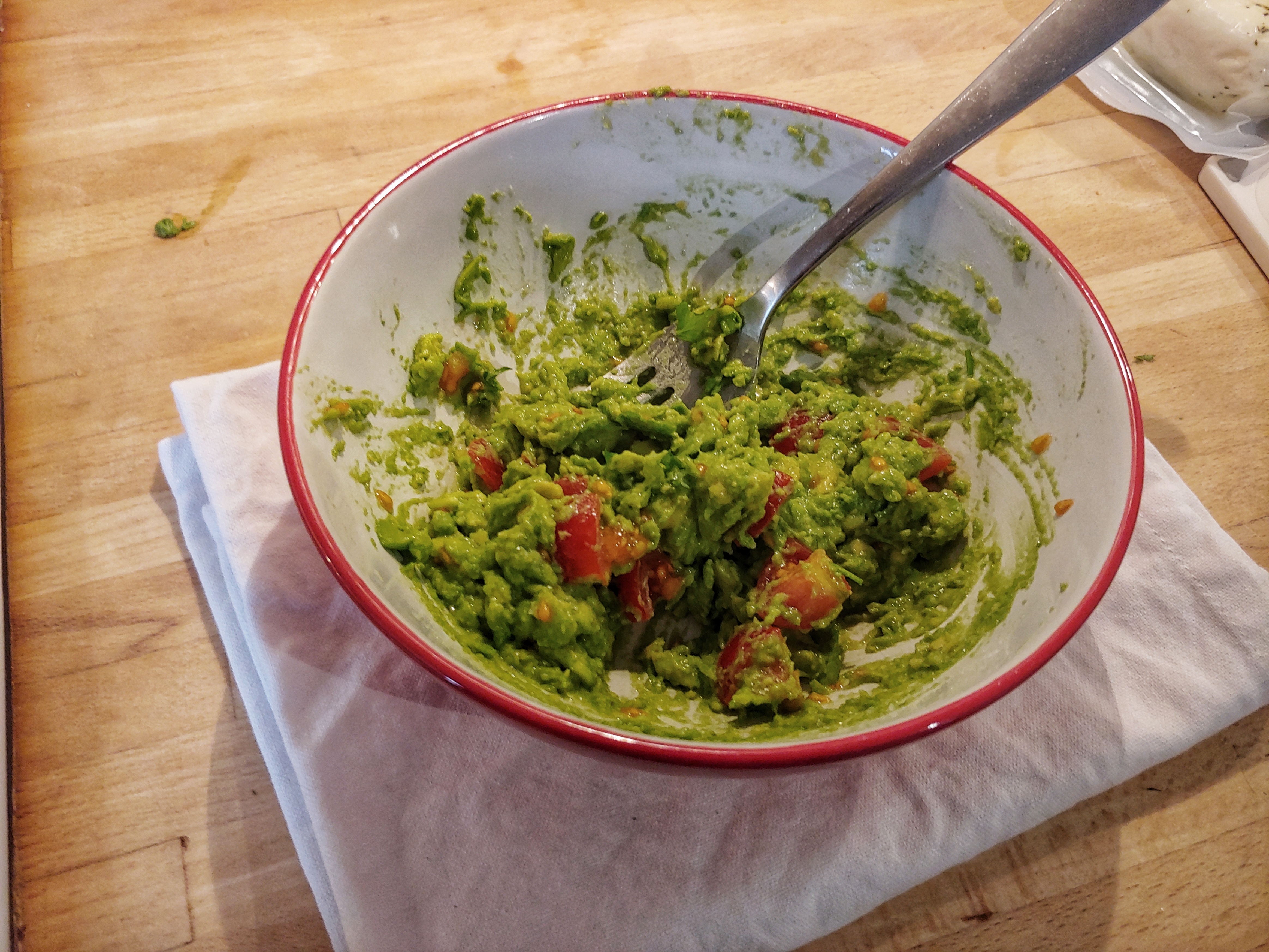 A white bowl with a red rim, with a fork sticking into guacamole with prominent pieces of red cherry tomatoes