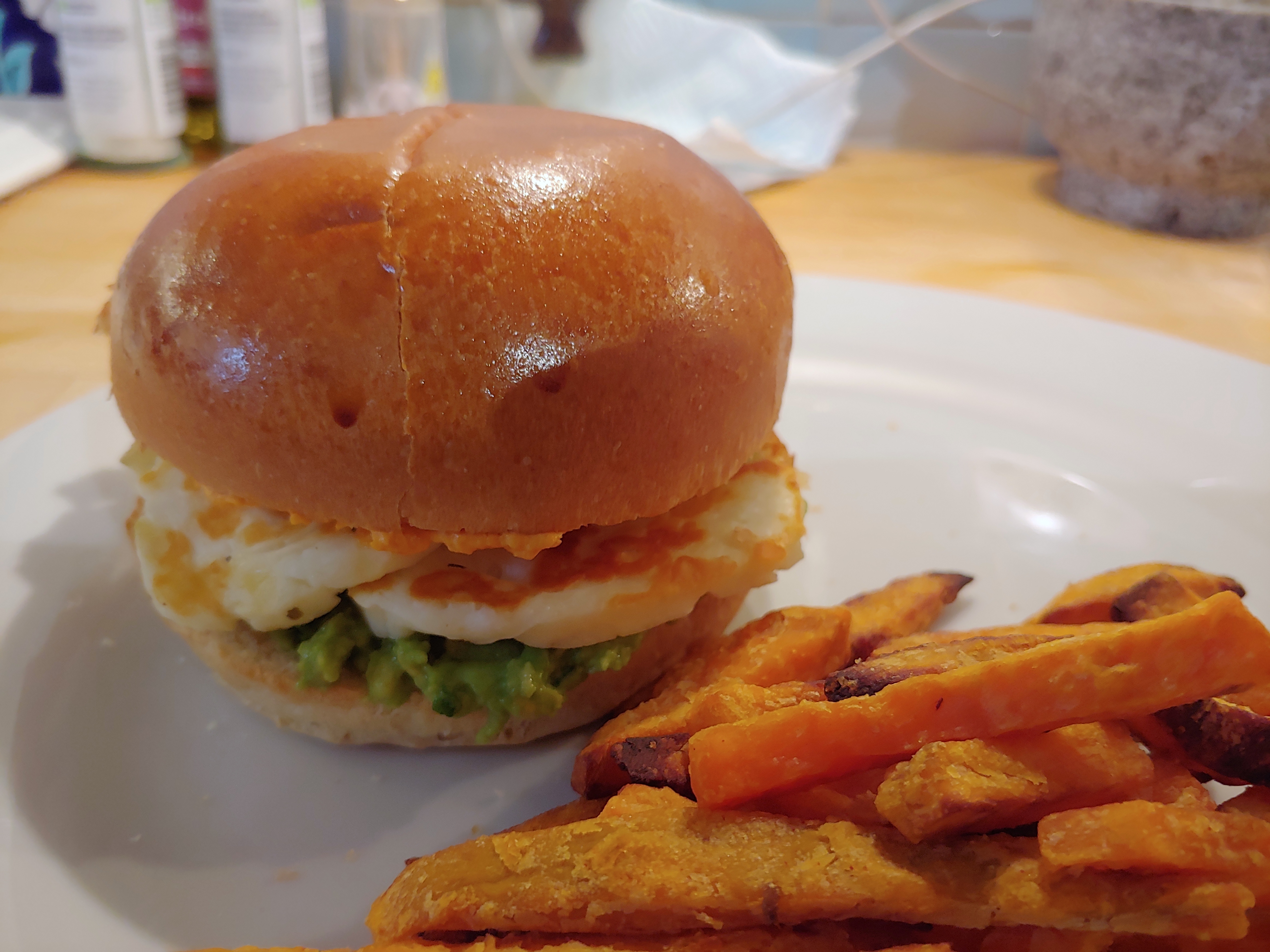 Halloumi, guacamole and houmous in a brioche burger on a white plate, next to a pile of sweet potato fries.