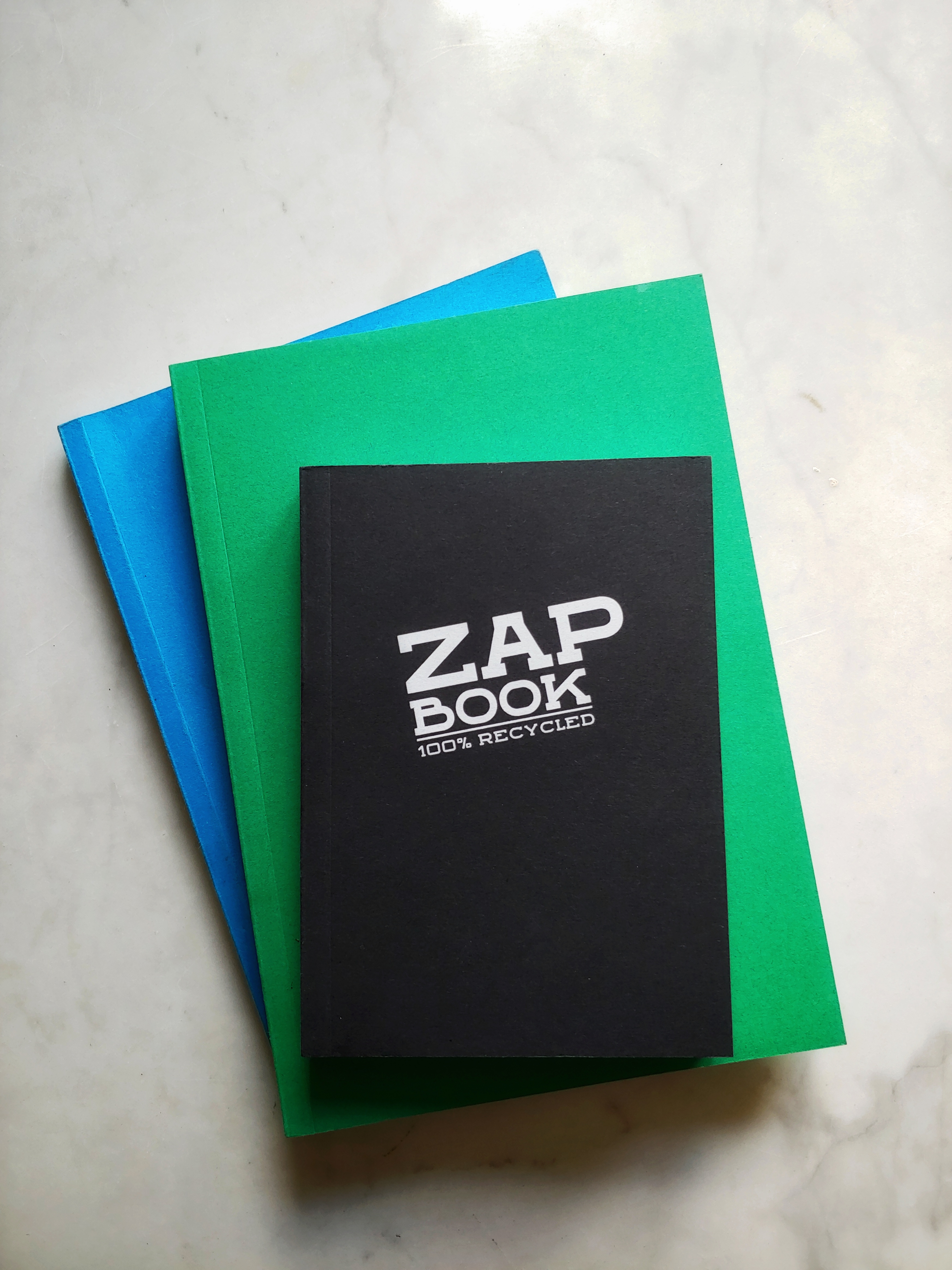 A pile of three Zap Notebooks, one blue and green in an A5 size, one smaller in black.  The black one reveals the cover, 'Zap Book 100% recycled in a bold modern font