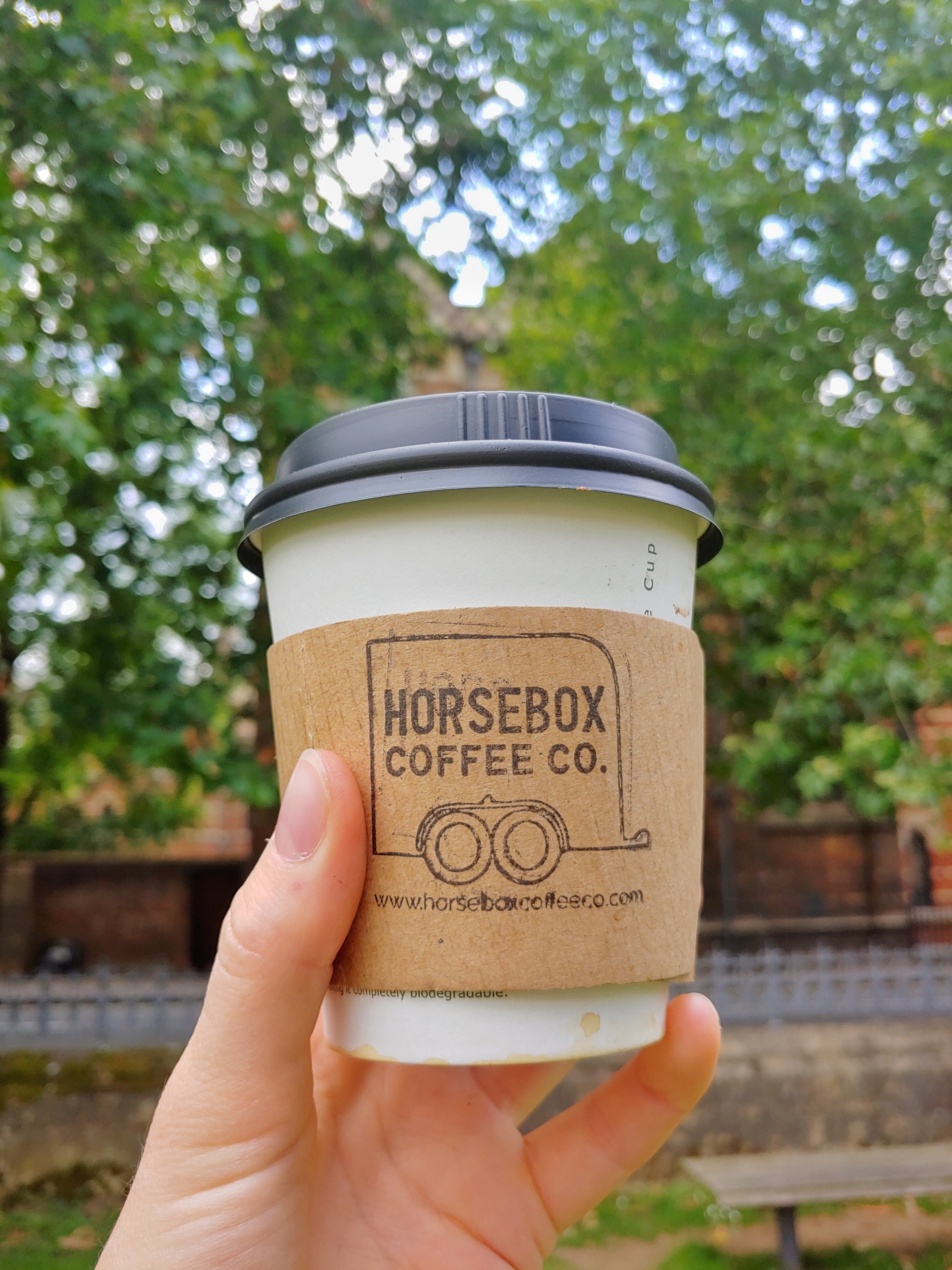 Image of coffee cup held up with trees and buildings of Keble College in the background.