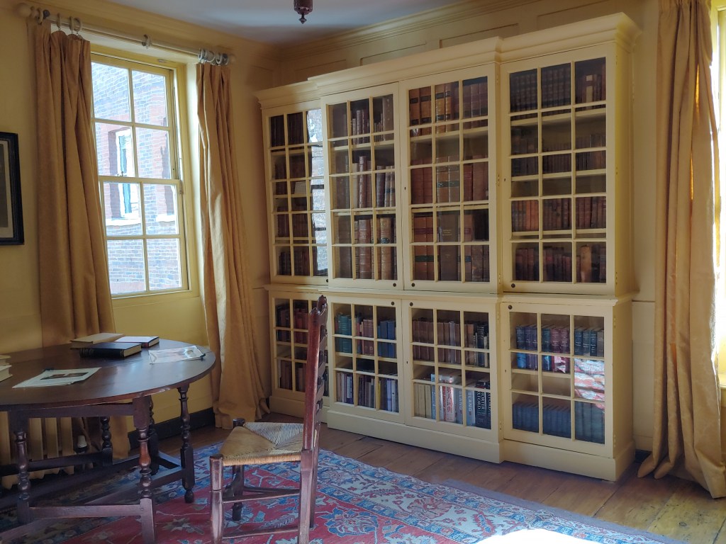 The room set up as a library, with a window in the corner, glass-fronted cupboard bookcases, and a small round table with chairs, where copies of books sit for visitors to read.