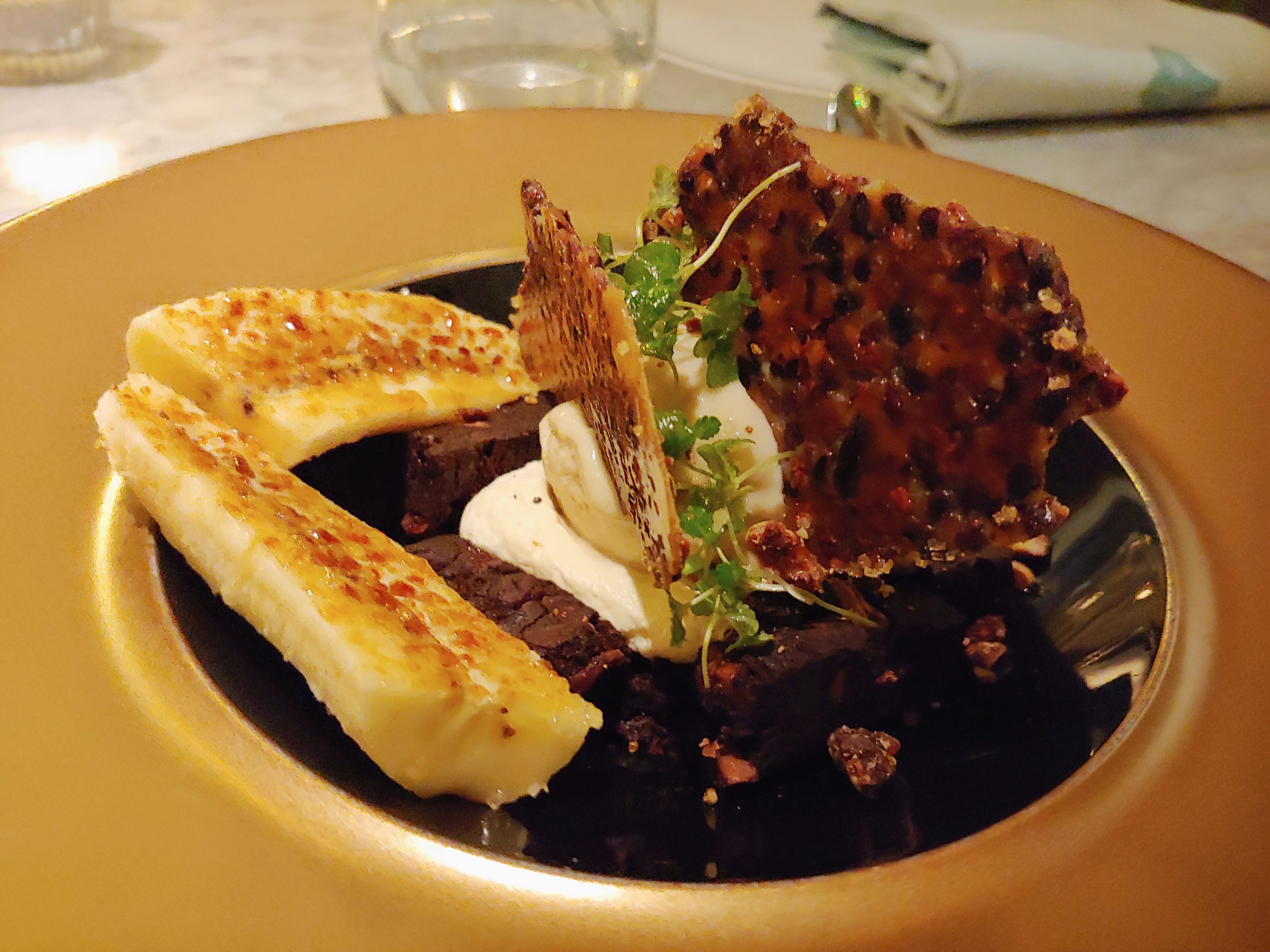 Image of a desert with a layer of chocolate brownie in a gold bowl, with two scoops of ice cream on top, each containing a chocolate nib tuille. Two lengthways slices of banana sit to the side with melted sugar on them.