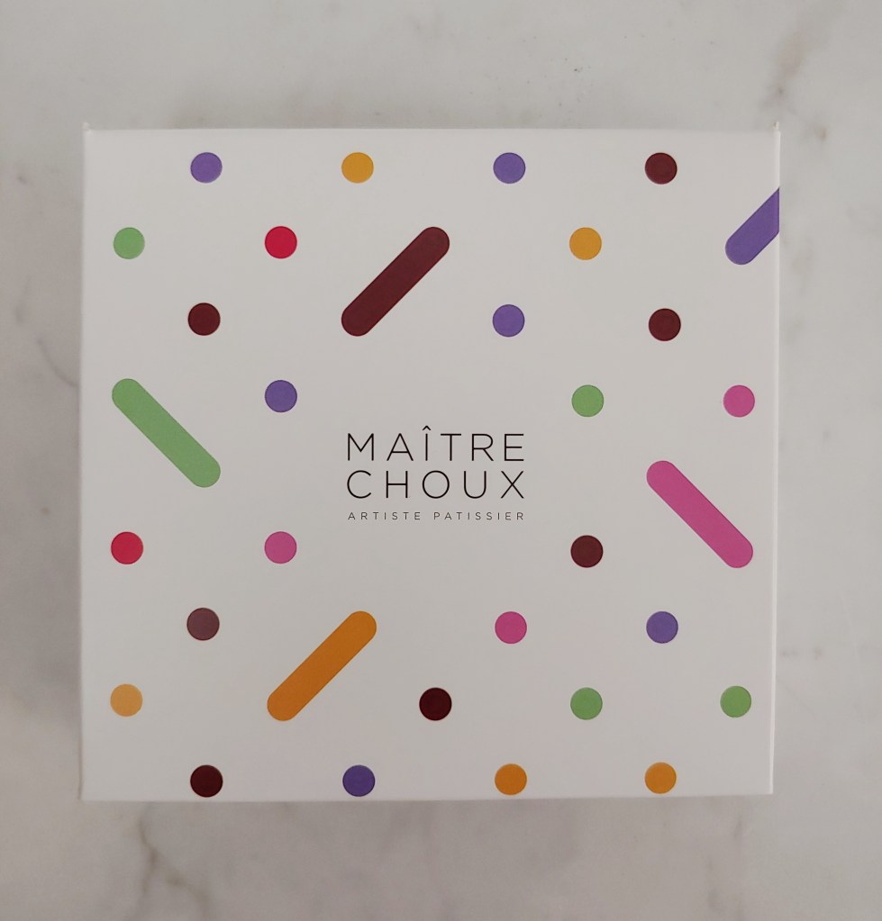Photograph of a square, white cardboard box with coloured dots and lines for decoration, and the words 'Maître Choux' in the middle.