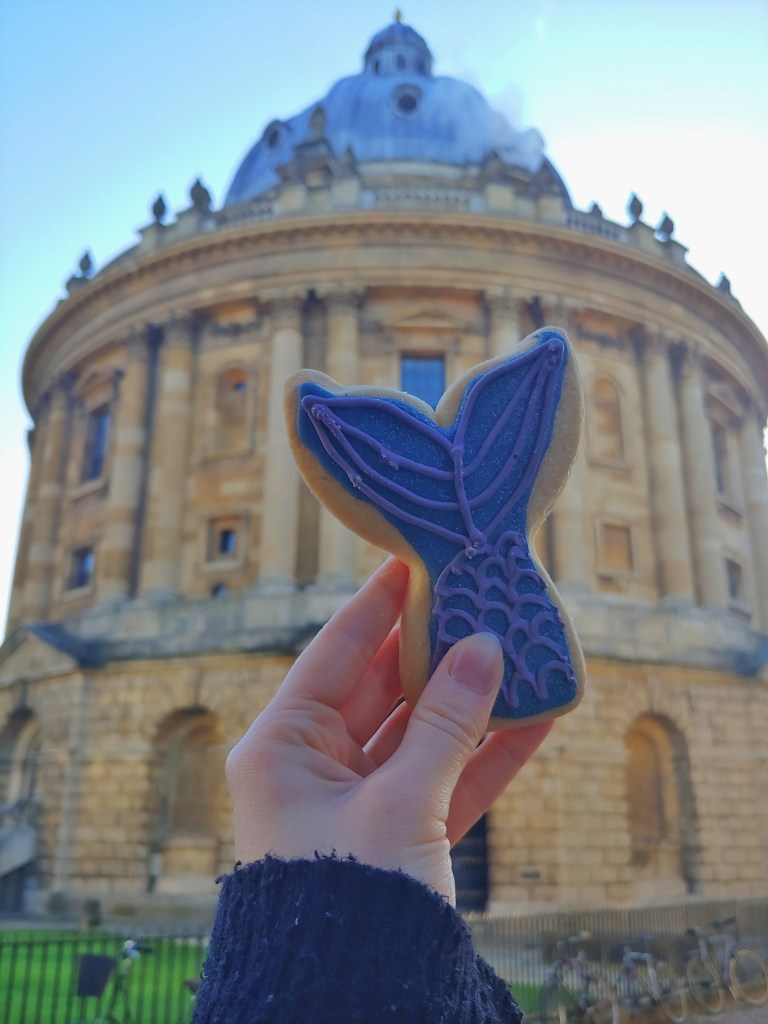 Photograph of a Mermaids Cookie being held up in front of the Radcliffe Camera, Oxford.  A woman's hand holds the cookie, which is in the shape of a mermaid tail, with blue glittery icing and purple piping.