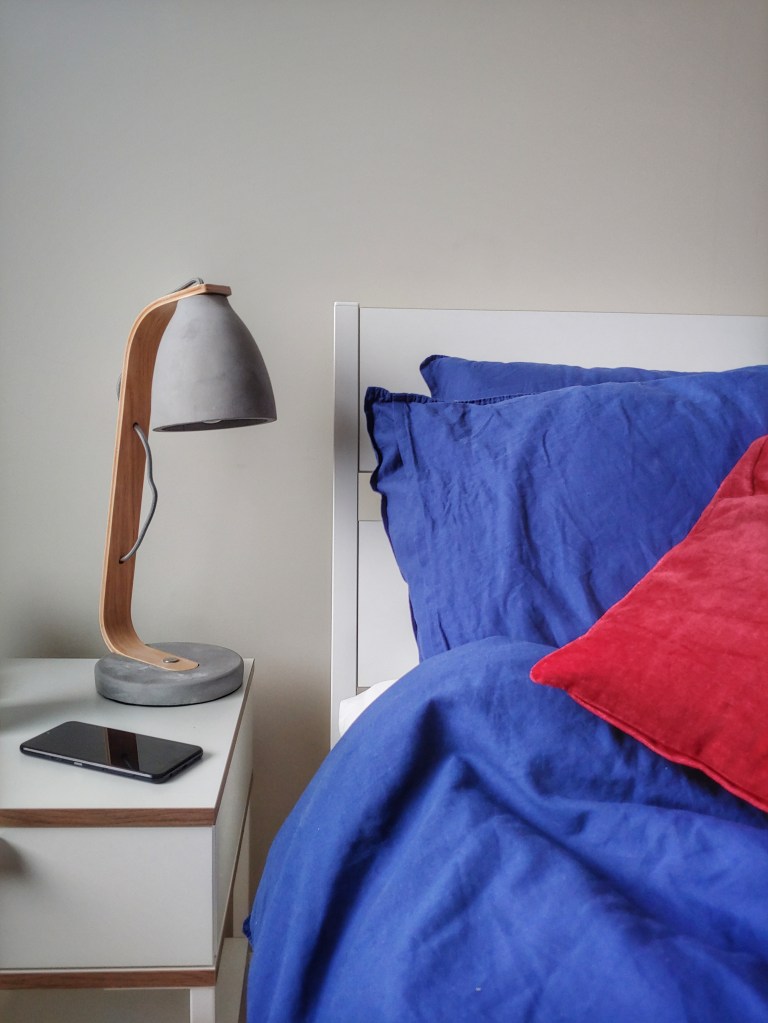 Close up of a bed with blue bedding and reddish pink cushion and a bedside table with a concrete lamp
