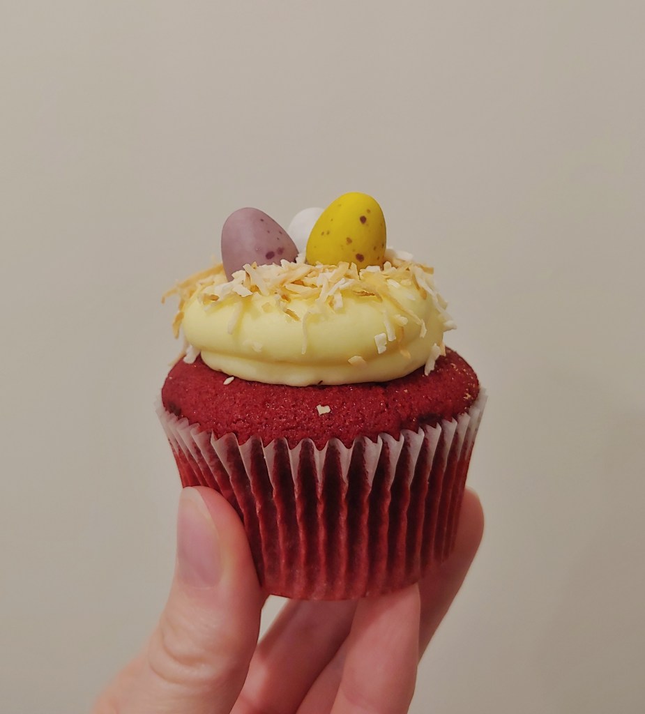 Close up of a red velvet cupcake with yellow buttercream icing with three chocolate eggs and coconut arranged to look like a nest.