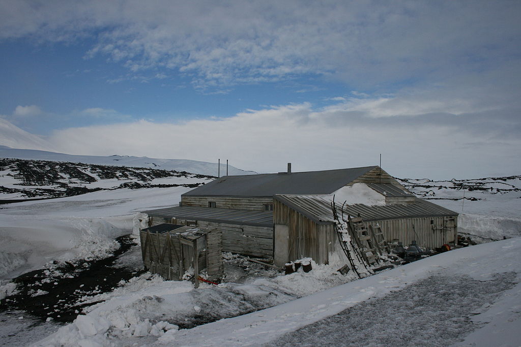 Image of Scott's Hut from the outside, with snowy hills in the distance, and a blue sky above.