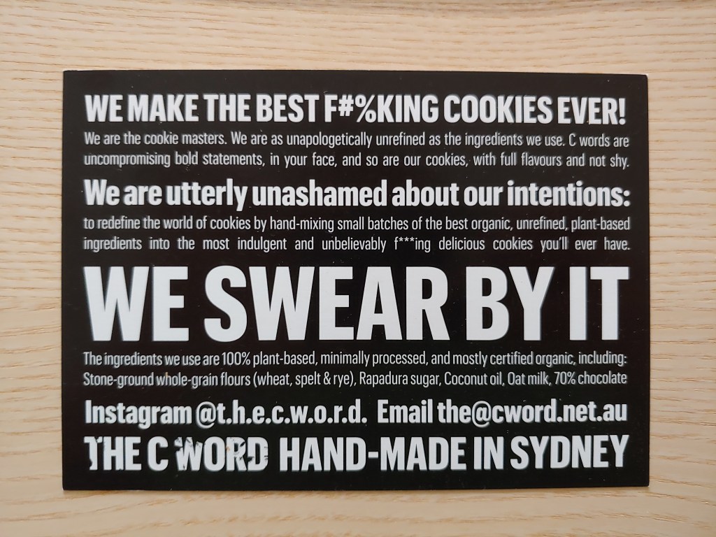 Postcard with white text on a black background reading 'WE MAKE THE BEST F#%KING COOKIE EVER!', 'WE are utterly unashamed about our intentions' and 'WE SWEAR BY IT' in bold standing out from other text about ingredients.