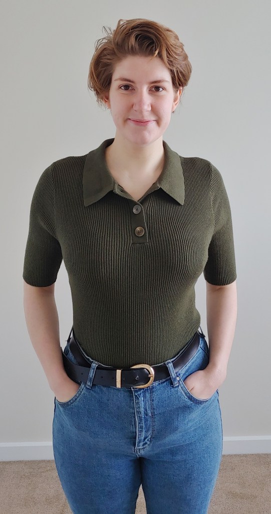 Photo of Helen wearing a dark khaki green ribbed knitted top with elbow length sleeves, a collar, and tortoiseshell buttons on the collar; the top is tucked into blue jeans.