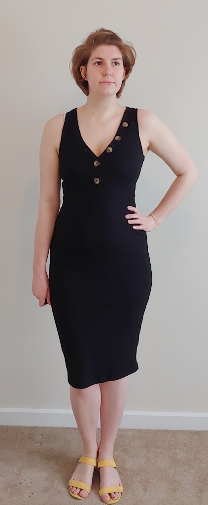 Full length photo of Helen wearing a black, ribbed sleeveless dress, with large buttons down a v-neckline, with yellow sandals.