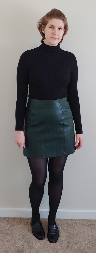 Full length photo of Helen wearing an above-the-knee A-line dark green faux leather skirt with a black ribbed turtle neck top and black flat pumps.