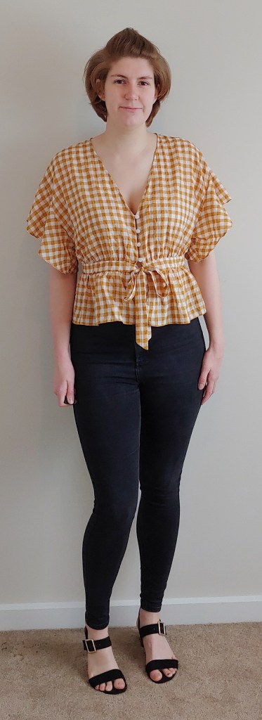 Full length photo of Helen wearing a yellow and white gingham top with floaty short sleeves and a peplum waist with tie, and black jeans.