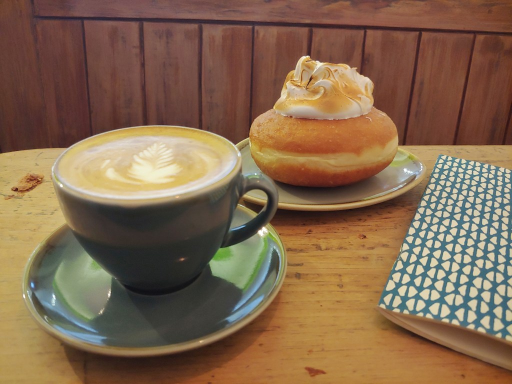 Blue coffee cup with froth, and a doughnut with a large meringue topping, on a wooden table next to a blue and white printed notebook.