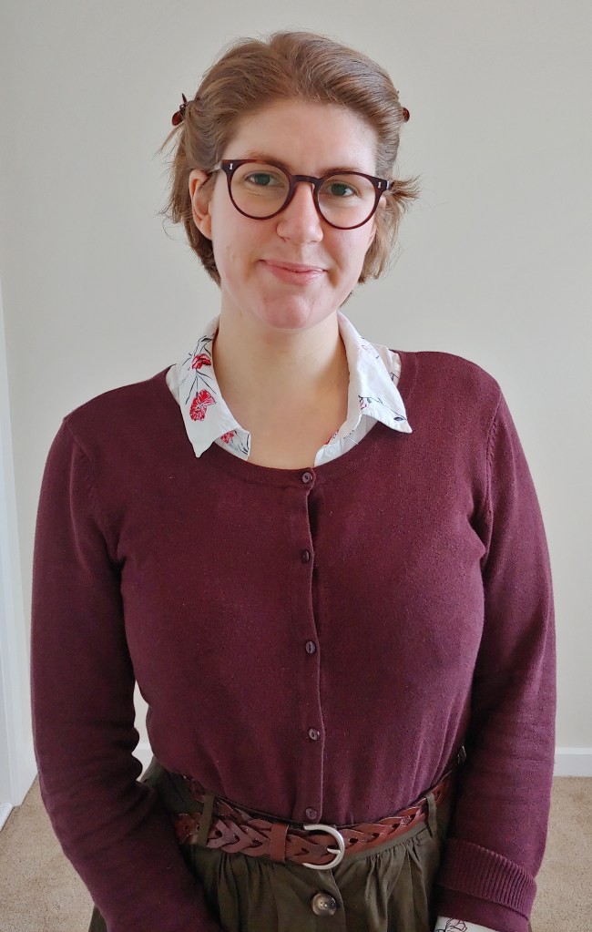 Head and shoulder shot of Helen wearing a burgundy cardigan with a white and floral print shirt underneath, and tortoiseshell colour round framed glasses.