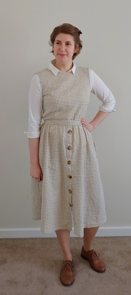 Full length photo of Helen wearing a light yellow/green and cream gingham dress over a white shirt with the sleeves rolled up, with brown brogues.