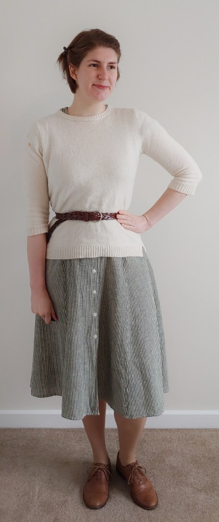 Full length photo of Helen wearing a green and thin white striped dress with a cream jumper over the top, with a brown belt at the waist and brown brogues.