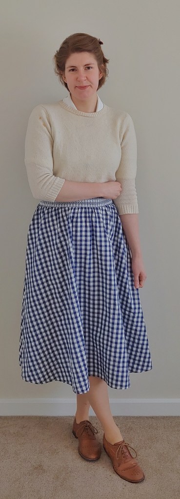 Full length photo of Helen wearing a cream half-length sleeved jumper over a cream shirt, with a blue and white gingham midi length circle skirt