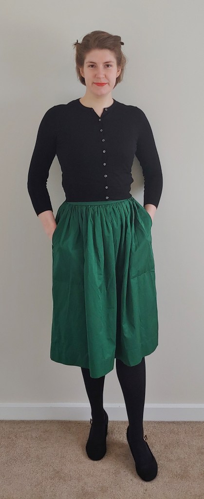 Full length photo of Helen wearing a dark green silky skirt with a cropped black cardigan, black tights and black Mary-Jane heels.