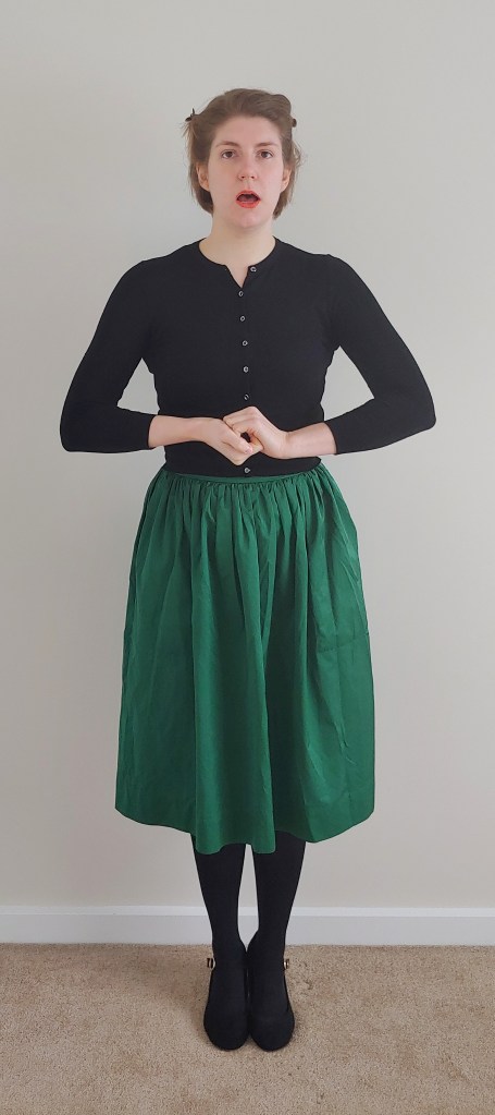 Full length photo of Helen wearing a dark green silky skirt with a cropped black cardigan, black tights and black Mary-Jane heels, stood with hands clasped in front of her and mouth open as if singing.