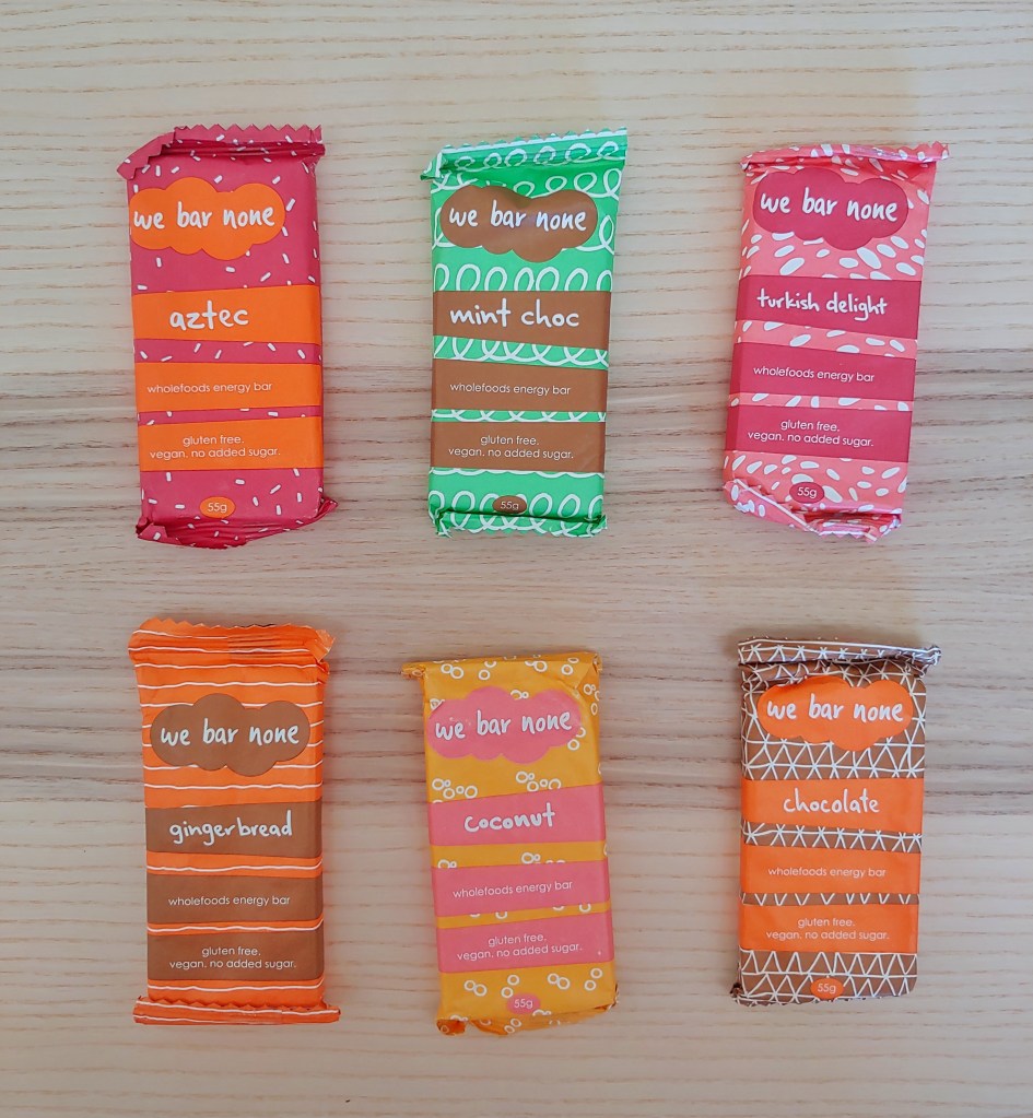 Top-down view of six wholefood bars of different flavours in colourful packaging on a wooden background.
