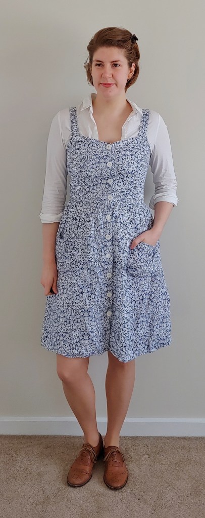 Full length photo of Helen wearing a knee-length pinafore dress with white buttons down the centre front, and patch pockets with a gathered skirt, in a white floral on blue pattern. Underneath is a simple collared white shirt with the sleeves rolled up to te elbows.