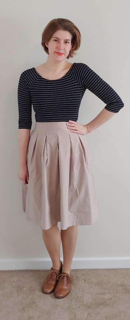 Full length photo of Helen wearing a beige knee-length pleated skirt with a black and thin white horizontal striped top with half-length sleeves