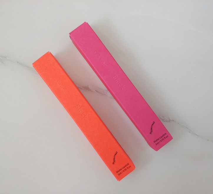 Two brightly coloured lipstick boxes, one pink and one orange-red, against a marble look background.