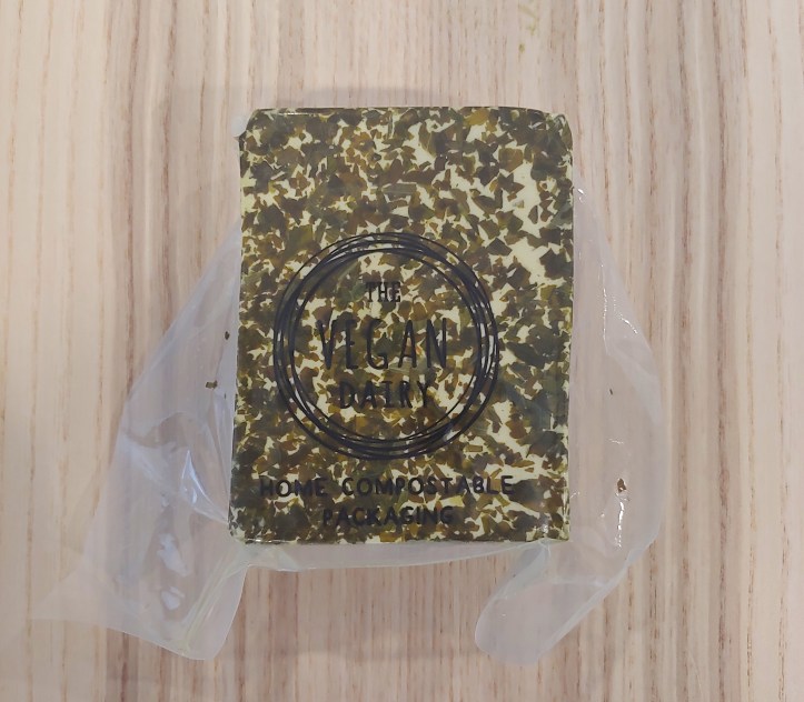Close-up of the a block of white cheese with green herbs coating the outside, in a clear package labeled 'Home Compostable Packaging'