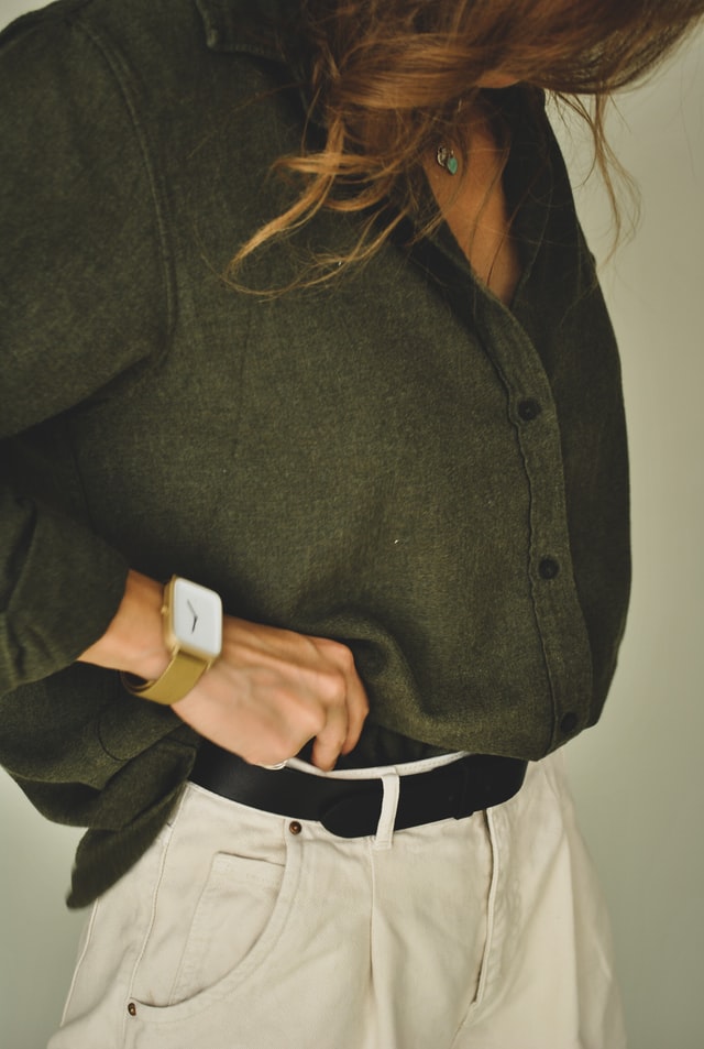 A woman with long brown hair wearing a large green shirt and cream chinos with a black belt. Close-up on the belt