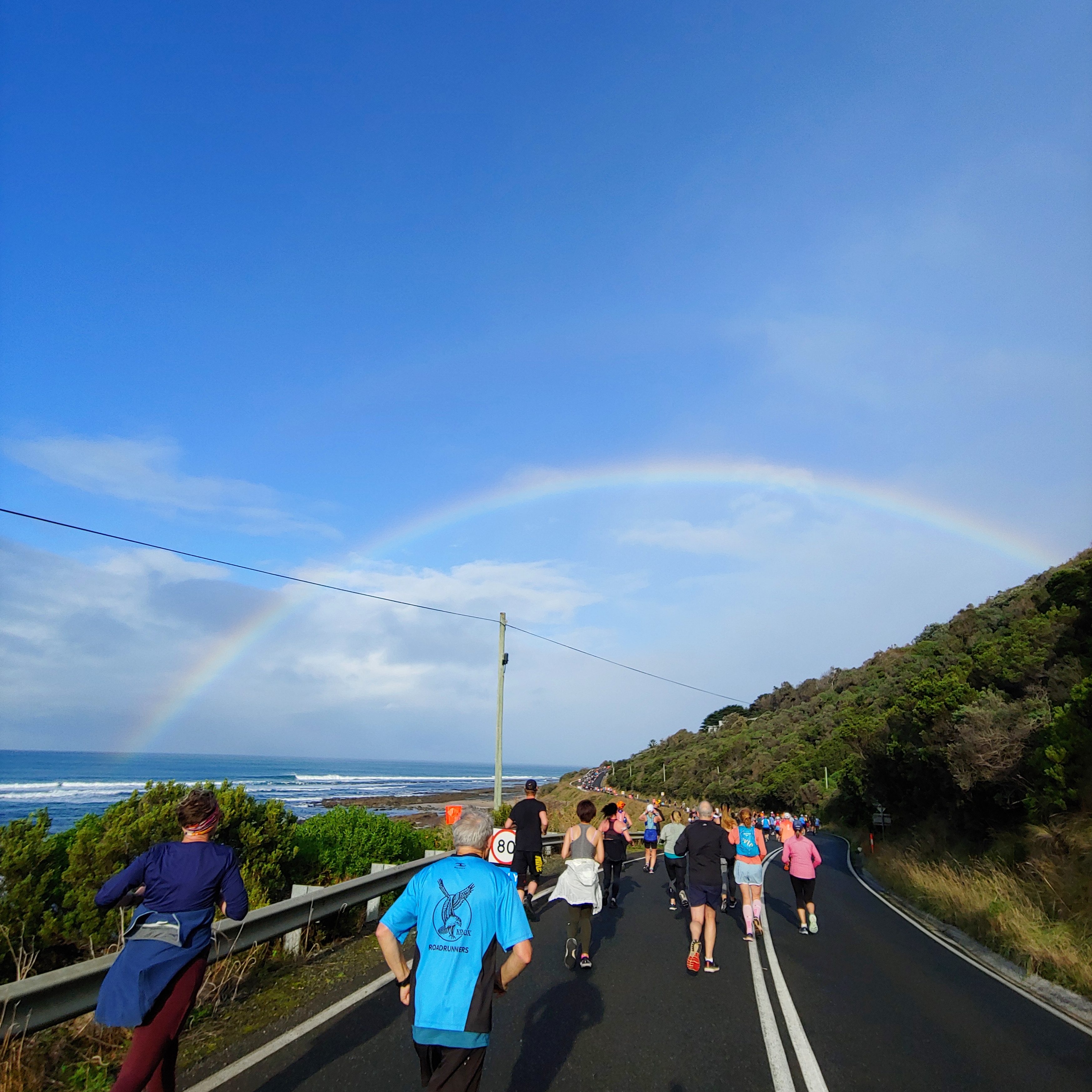 People running along a tarmac road with woods on the right and the sea on the left, and a bright rainbow in the sky overhead.