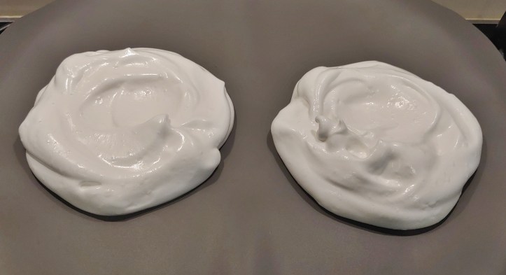 Two nearly circular white mounds of vegan meringue on a gray baking tray waiting to be cooked.