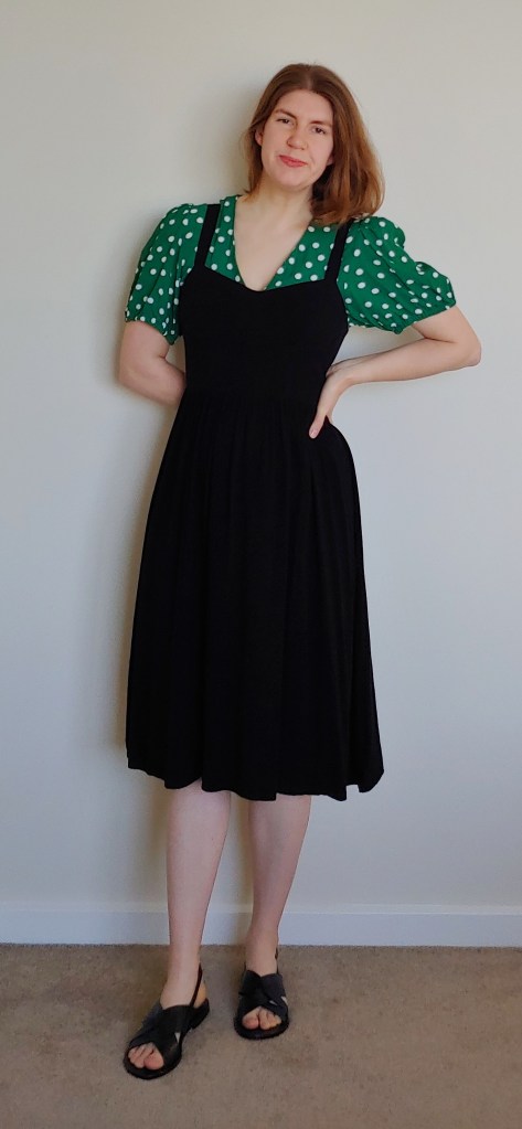 Helen wears a grass-green puff-sleeved shirt with medium-sized white polka dots, and a black pinafore dress, with black sandals.