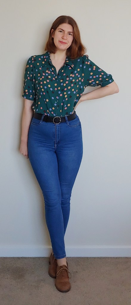 Helen wears blue skinny jeans and a teal-green blouse with medium-sized polka dots in dark blue, light blue, pink and beige. She also wears brown brogues.