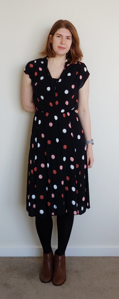 Helen wearing a black mid-length dress with medium-sized pink, coral pink and white polka dots. She also wears black tights and brown ankle boots.