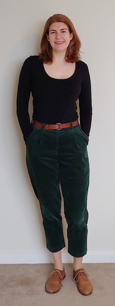 Full-length photo of Helen wearing forest-green corduroy trousers with a long-sleeved, scoop neck t-shirt, a thin brown belt, and brown brogues.