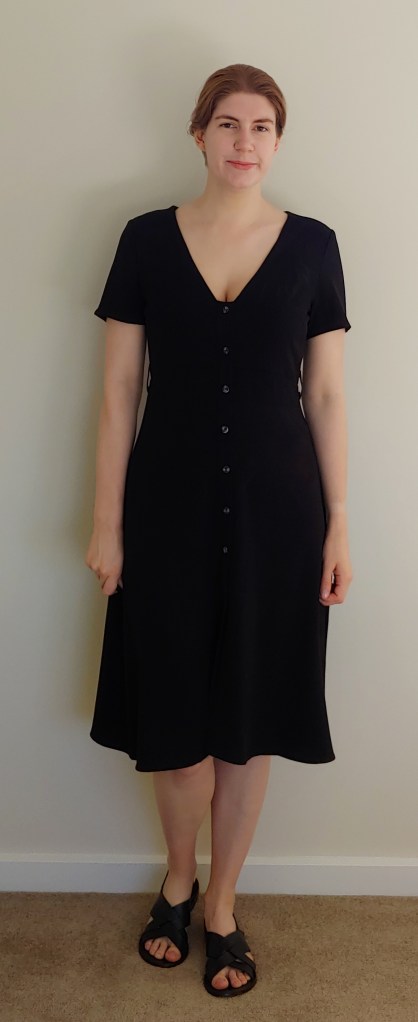 Full-length photo of Helen wearing a knee-length black A-line dress with buttons up the front and short sleeves, with a pair of black criss-cross sandals.