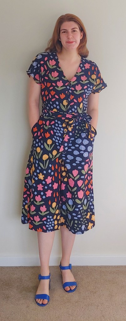 Full-length photo of Helen wearing a floral wide-leg, mid-length hump suit with a v-neck and short sleeves, with blue block-heel sandals.