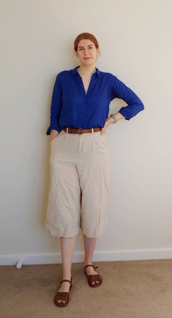 Full-length photo of Helen wearing calf-length cream-coloured culottes with a dark blue linen shirt with sleeves slightly rolled up, a thin brown belt and brown, rounded flat sandals.