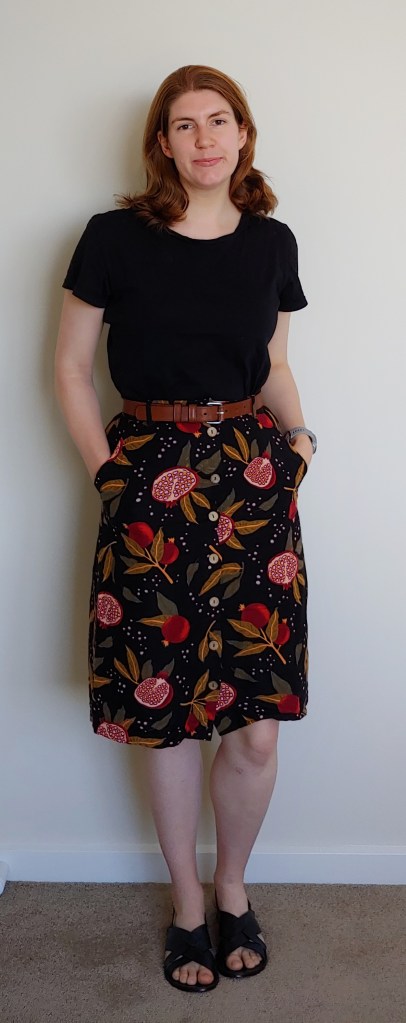 Full-length photo of Helen wearing a black t-shirt with an A-line skirt printed with red pomegranates and brown and green leaves on a black background, with criss-cross black sandals.