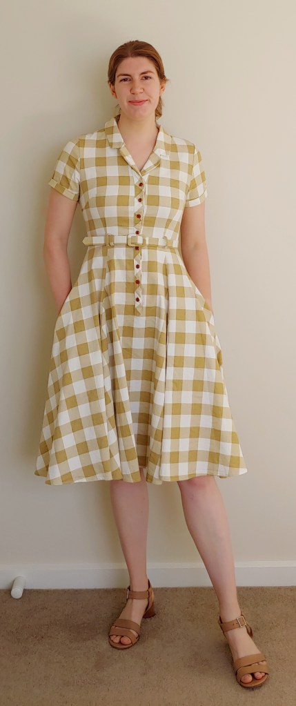 Full-length photo of Helen wearing a short-sleeved, knee-length yellow and white gingham dress with full circle skirt, buttons to the waist, small lapels and a matching belt, with light-brown block heel sandals.