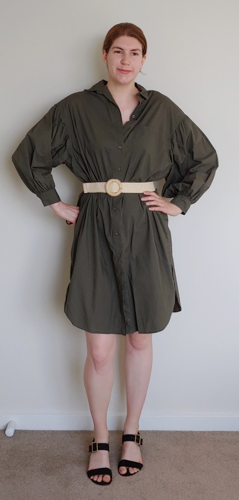 Full-length photo of Helen wearing a khaki green shirt-dress with long, puffy sleeves, with a straw belt with round buckle, and black block-heel sandals.