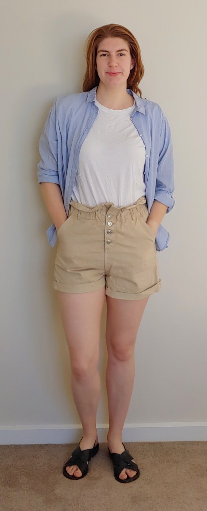 Helen wearing high-waisted beige shorts with visible button-fly and paperbag waist, with a white t-shirt and light blue shirt open over the top. She also wear black crossover flat sandals.