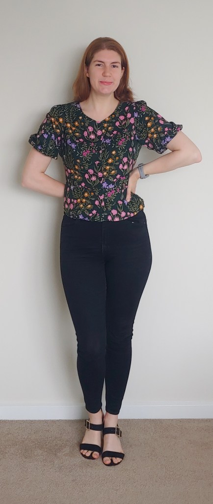 Helen wearing a short puff-sleeved fitted blouse in a dark green colour with pink, yellow, and purple floral print, and black Levi's high-waisted skinny jeans, with black block-heeled sandals.