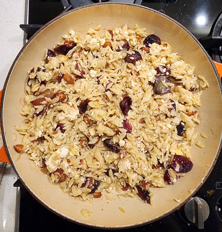 Top-down image of orzo, grapes, feta and almonds mixed together in a round, orange Le Creuset roasting dish.