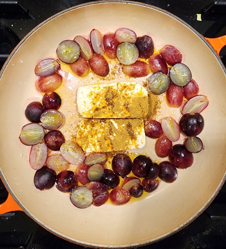 Top-down view of the roasting dish, with feta coated in ground cumin in the centre, surrounded by halved grapes.