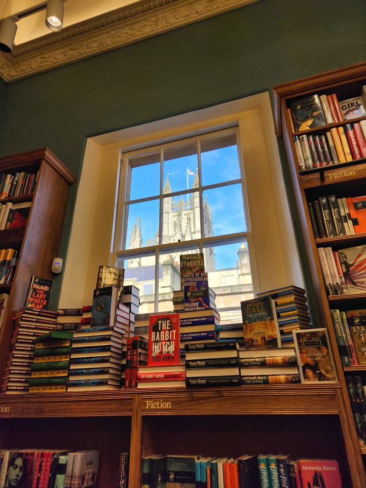 View from inside looking across stacks of books to the window and a view of the tower of Bath Abbey.