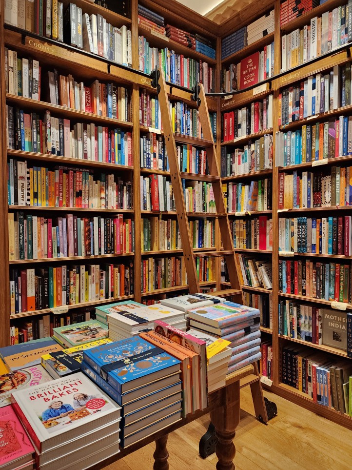 Corner bookselves stuffed with books with a shelf ladder, and a table piled high with books in the foreground. 