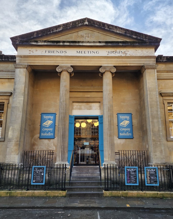 Classical style building with large pediment and Ionic columns, with a large doorway with blue doors on either side, and two notice boards reading 'Topping & Company Booksellers' in gold text on a light blue background. 'Friends Meeting House' is written in classical capital letters beneath the pediment.