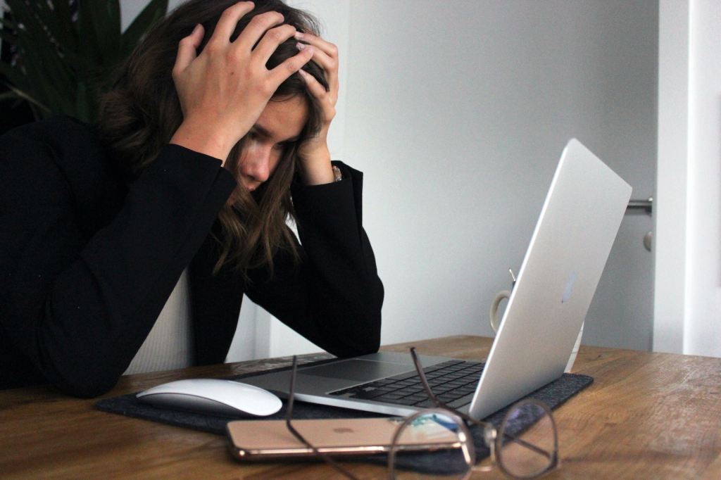 Woman at a desk in holding her head in her hands looking at a laptop screen.
