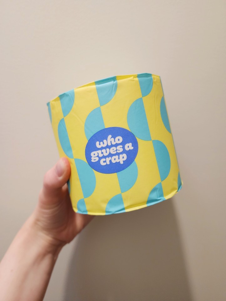 Hand holding a roll of toilet paper packed in turquoise and yellow patterned packaging.